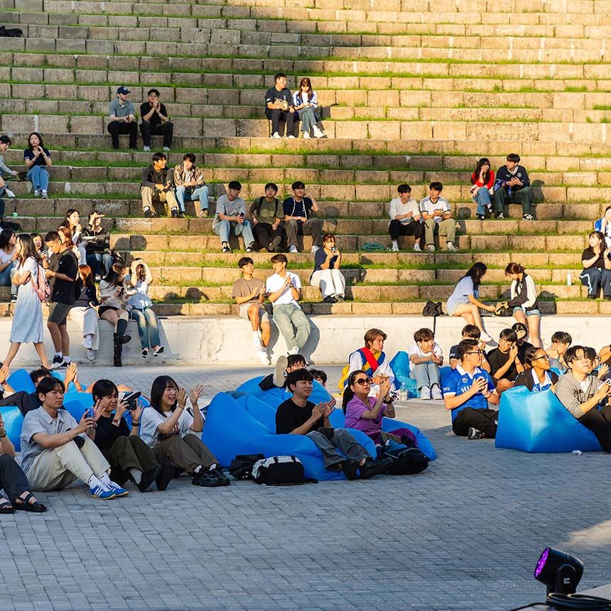 A group of students sits on stone amphitheater steps and blue inflatable loungers during an outdoor campus event.