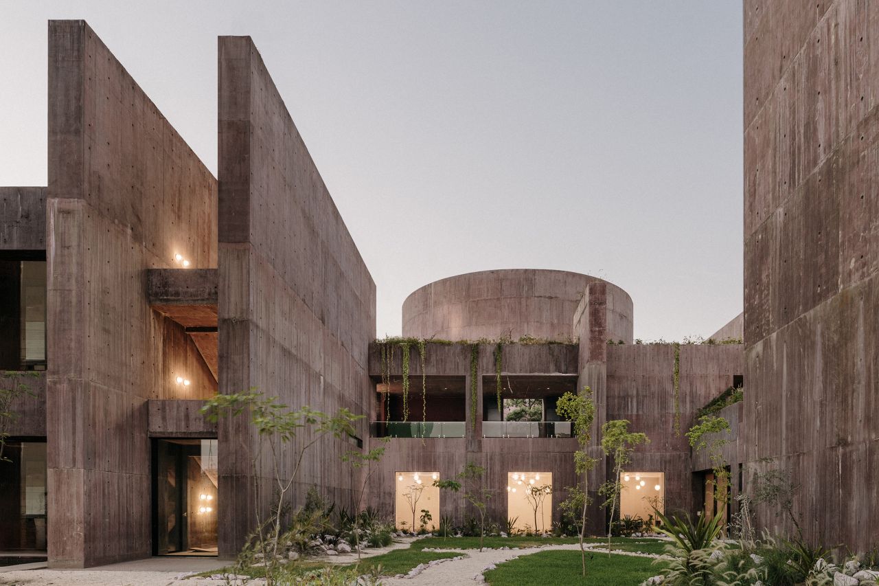 The exterior of a modern building made of concrete with tall, imposing wall and a cylindrical structure in the background. In the foreground is a landscaped courtyard.