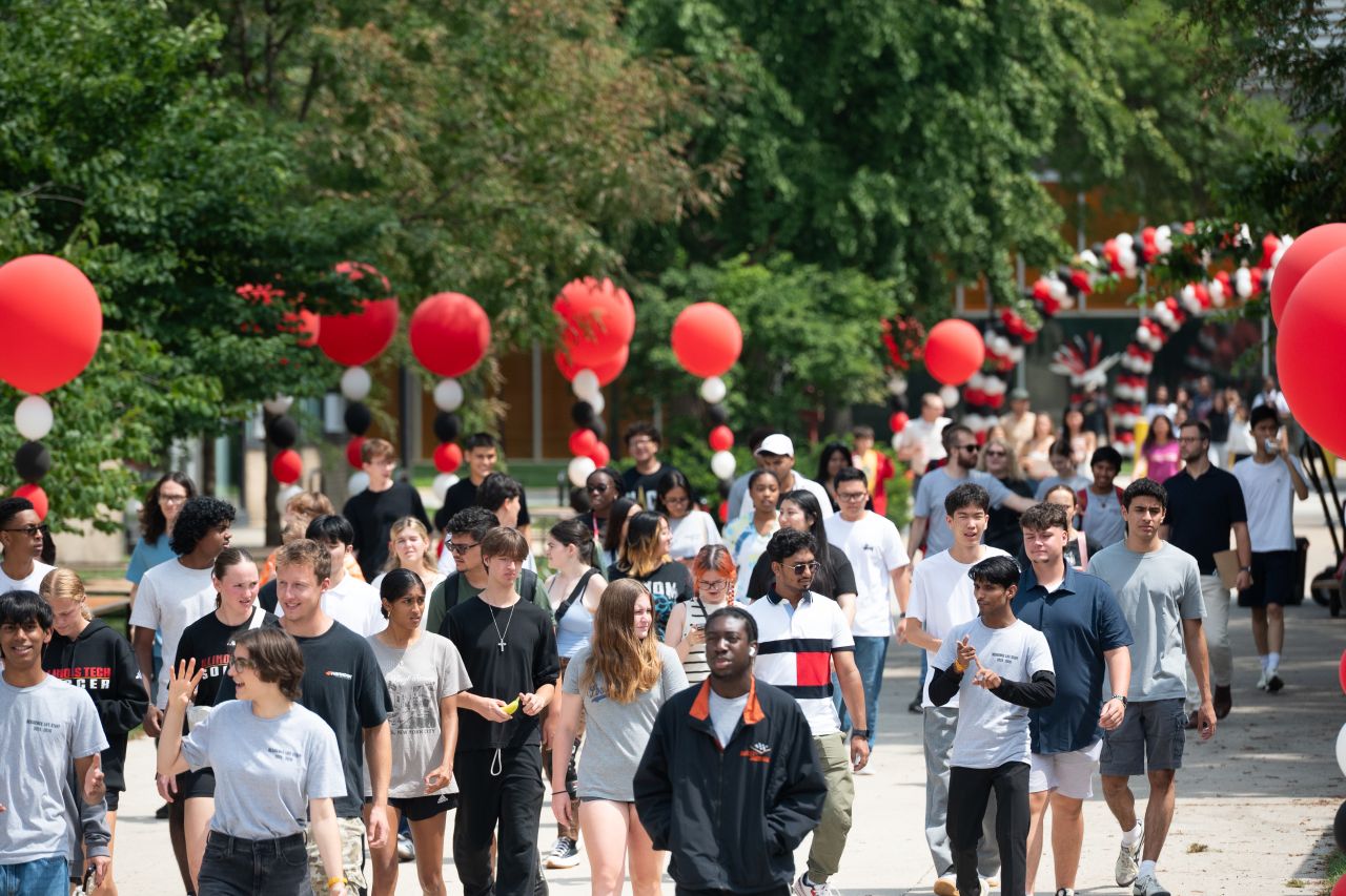 A large group of college students walks on a sidewalk outside. There are several red, black and white balloons decorating the area. There are many trees and a building in the background.