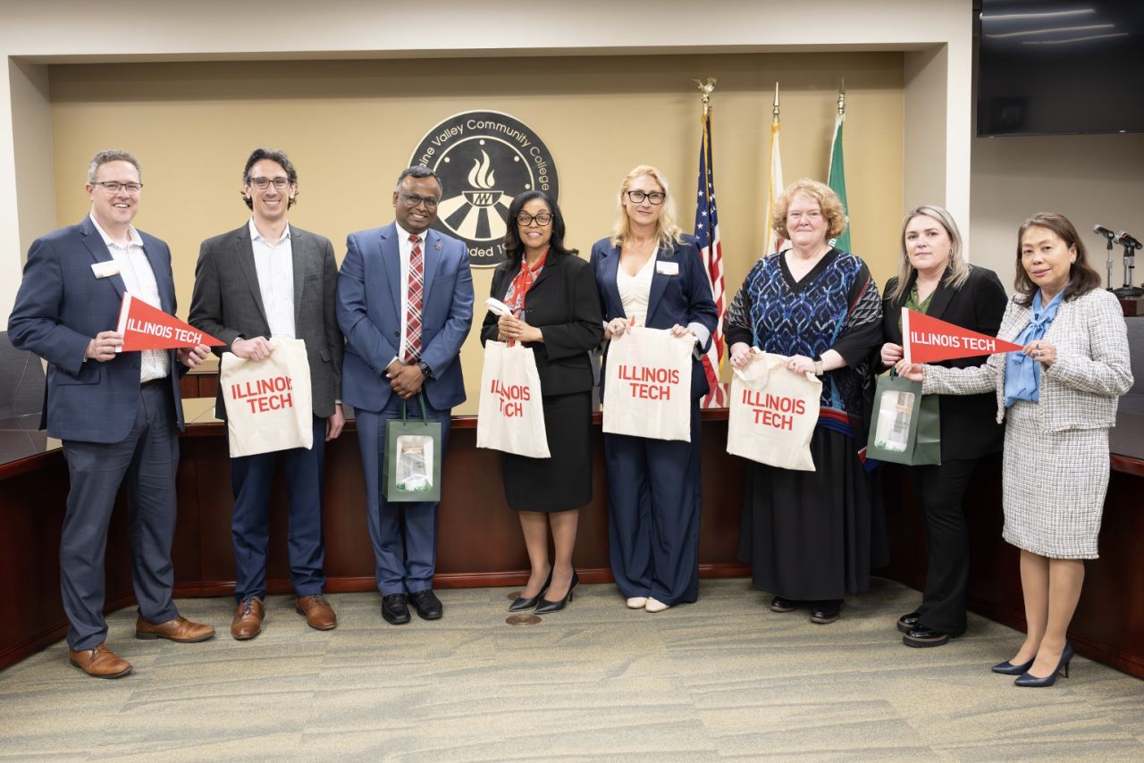 Eight people pose for a photo in front of the seal of Moraine Valley Community College. Several of them are holding pennants and bags with the words Illinois Tech.