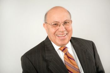 A man in a dark suit, white shirt and striped orange tie smiles for a portrait in front of a neutral background.